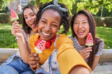 Three happy young women are sharing watermelon ice cream, smiling for a selfie. Their faces glow...