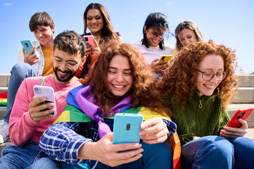 Smiling young friends using mobile phone celebrating gay pride festival day together. Happy LGBT people community with rainbows flags watching and checking social networks outdoor