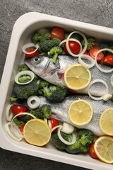 Raw fish with vegetables and lemon in baking dish on grey table, top view