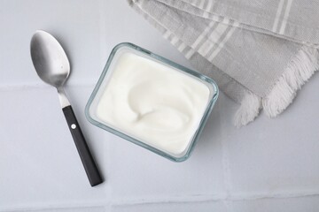 Delicious natural yogurt in glass bowl and spoon on white tiled table, top view
