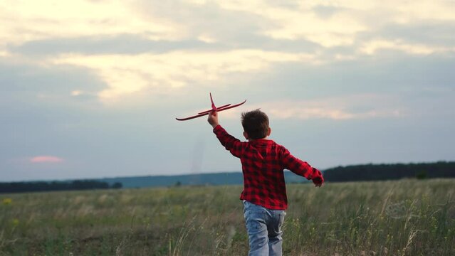 Casual playful boy kid running with red plane pilot flying at evening field dramatic sky back view slowmo. Adorable male child playing airplane flight fantasy imagination enjoy freedom happy childhood