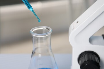 Laboratory analysis. Dripping blue liquid into flask near microscope on table, closeup