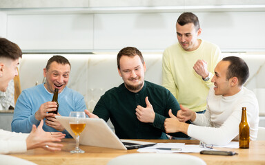 Fototapeta premium Group of men watching sports match online on laptop during friendly meeting with beer at home, excitedly celebrating winning score in game ..