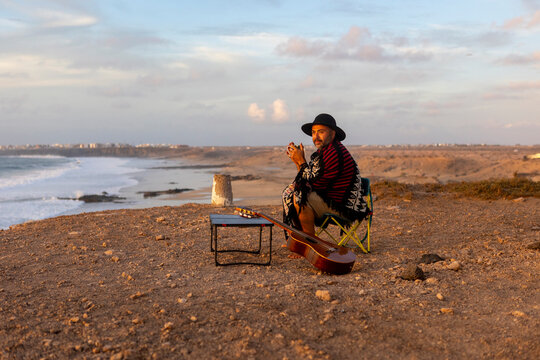 Nomadic Musician drinking hot drink and Composing by the Seashore at Sunset