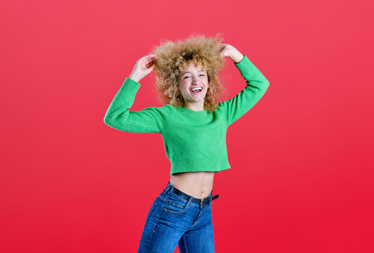 A woman with voluminous curly hair enjoys a playful moment, smiling and touching her hair, clad in a vibrant green sweater and blue jeans, set against a vivid red backdrop