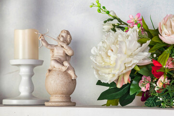 White statue of cupid boy with bow and arrow on shelf next to candle, close-up. Small statue amur near bouquet of flowers. Concept of calm and serenity, peaceful atmosphere. Copy ad text space poster - obrazy, fototapety, plakaty