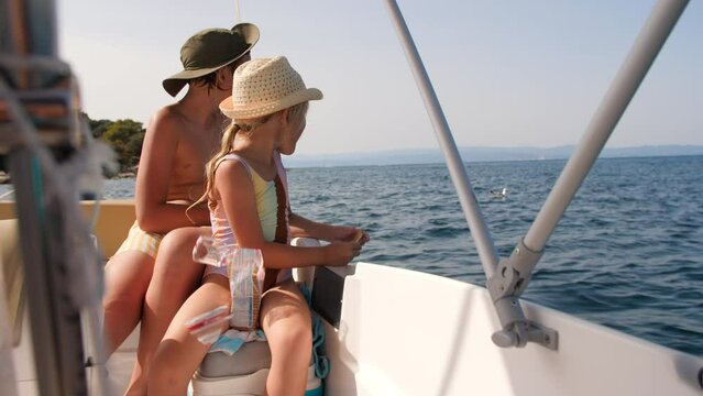 A family on a boat trip, a young girl and a teenage brother, both wearing hats, feed cookies to a seagull that floats nearby