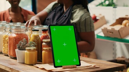 Tablet with greenscreen display placed on farmers market counter, next to homemade additives free sauces and cooking products in recyclable jars. Merchant displaying blank mockup template.