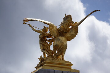 Shiny gold statue of pegasus and person under bright blue sky and clouds