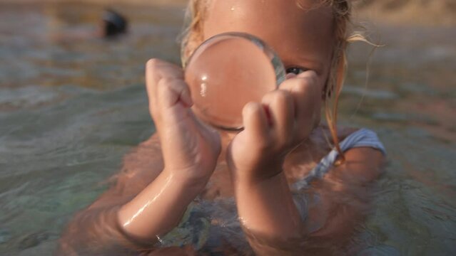A girl, partially submerged in sea water, holds a glass ball in her hands, looking through it with concentration with a thoughtful expression on her face