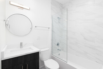 A modern bathroom with a dark wood cabinet, circular mirror above a white marble countertop, and a shower with large marble tiles.