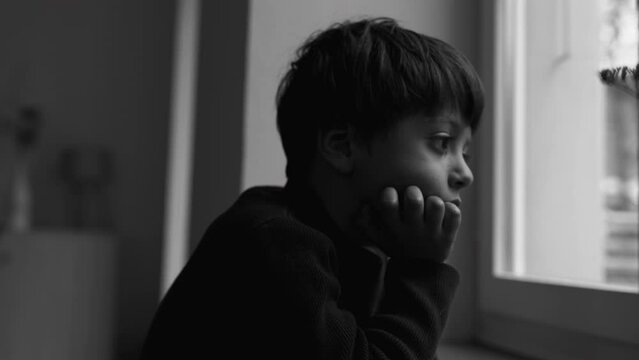 Depressed child stands by window gazing at view with introspective sad expression in moody monochrome, dramatic black and white. kid feeling solitude