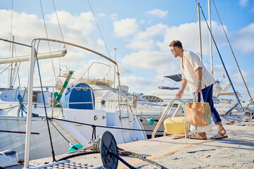 Adult man on casual clothes boarding a sailboat carrying a coolbox and bags at the yacht marina....