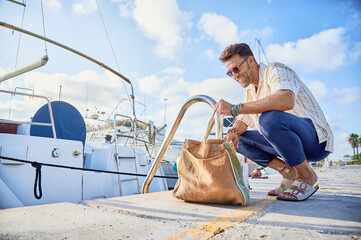 40s wealthy man ready to board the docked sailboat checks the bags with provisions at the pier of...
