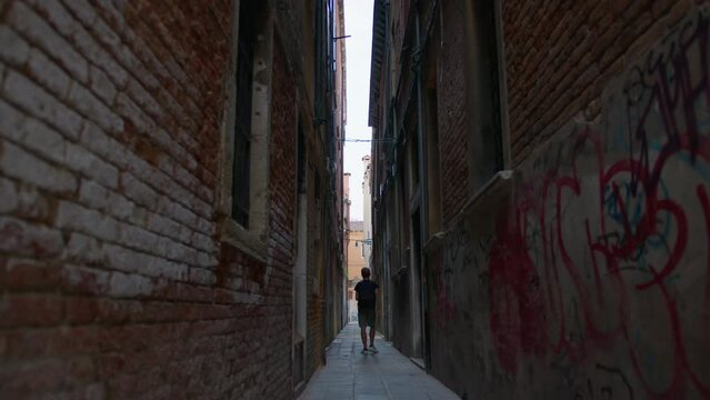The man walks away down a narrow alley lined with brick walls and painted with graffiti. Venice, Italy