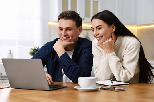 Happy couple using laptop together at wooden table in kitchen - Powered by Adobe