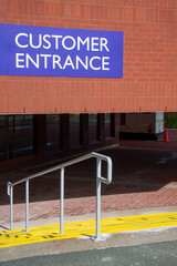 The exterior of a brown brick office building with a bright blue colored sign. The text, customer entrance, is in white letters. There are steps with a metal handrail down to a covered entrance.  