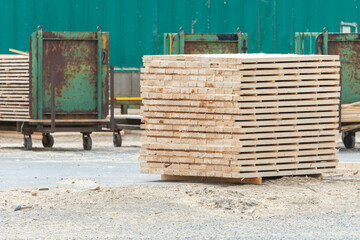 A lumberyard of a hardware store with bulk stacks of short-cut lumber, boards, and rough planks stockpiled for sale. Behind the stacked materials are trolly lumber carts and a green metal wall. 
