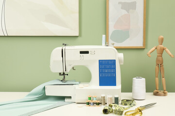 Modern sewing machine with cloth and craft accessories on white table near light green wall
