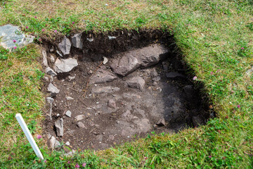 Small square shaped hole dug for exploration as part of an archeology dig. The ground is green grass with topsoil and rocks beneath. There's a white marker in the corner of the excavated artifact area