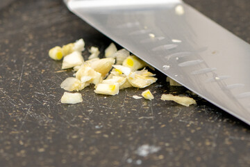 A stainless steel chef knife crushing, grating, cutting, grating, mincing, and chopping organic white garlic on a grey plastic cutting board. The root vegetable of an herb has been pressed to release 