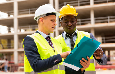 Two civil engineers working at the construction site are discussing the construction plan, holding documents in their hands and carefully studying them