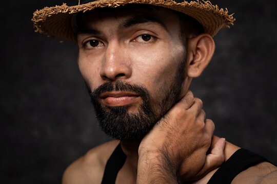 Portrait of Asian bearded man in hat standing in dark room against a black background and looking at camera.