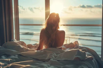 Fototapeten Woman on bed looking out the window overlooking the sea and sunrise © InfiniteStudio