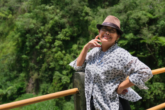 A woman wearing a hat leaning against the railing of a bridge, green trees in the background; smiling, happy expression