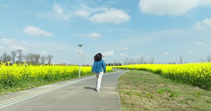 back view of happy young woman walking on the countryside road at spring sunny day with blooming oilseed flower field