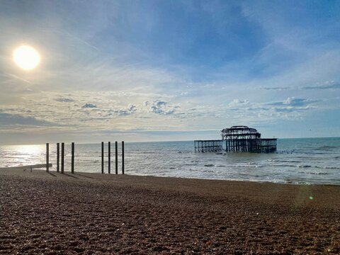 Ruin of burnt our West Pier off pebbly Brighton beach with blue sky and calm seas at low tide 