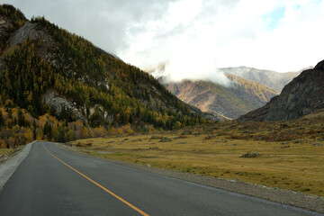A two-lane asphalt road turns into a wide crevice towards high mountains with peaks in the clouds on a sunny autumn day.