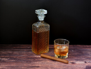 A crystal decanter, a glass of old whiskey with ice and a Cuban cigar on a wooden table.