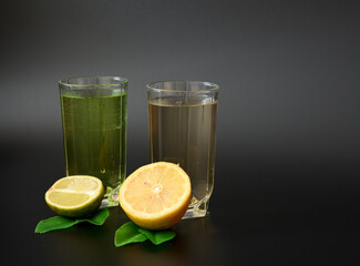 Two glasses of citrus juices on a black background, next to pieces of ripe lemon and lime with...