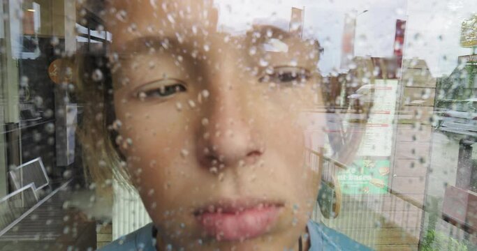 The boy looks thoughtfully through a glass window, speckled with raindrops, with a reflection of the street, a cafe window