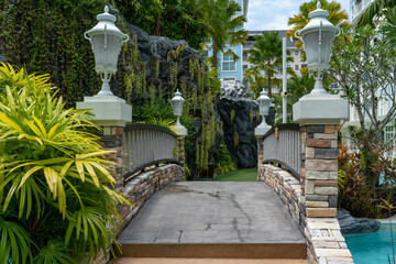 A stone pedestrian bridge with decorative lanterns and steps spans the blue water pool of a condominium at a tropical resort. Lush tropical vegetation grows around.