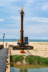 An old excavator with a long boom standing on the sandy shore of the sea next to a bridge spanning a river. A woman in a large hat stands on the bridge and looks into the distance.