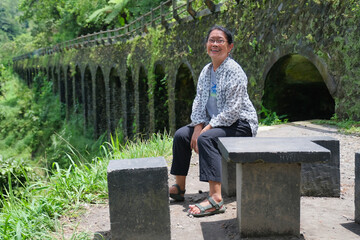 A woman sitting on a concrete bench with the pillars of an old brigde in the background; smiling,...