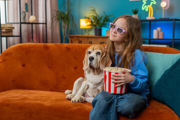 Excited young girl in 3D glasses eating popcorn and watching interesting TV serial, sport game, film, online movie content. Preteen child kid sits beside beagle dog on sofa in living room at home.