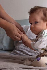 Bebé de ojos azules y cabello castaño sentado de costado en su cama agarrando las manos de su mamá en su habitación