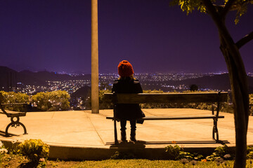 Mujer de espalda usando un beani rojo sentada en una banca de un parque iluminada con la luz amarillo de un poste en un parque de noche de invierno en la cima de la ciudad