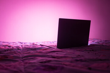 Laptop abierta encendida iluminando la pared blanca de una habitación, está sobre una cama y la luz es morada.