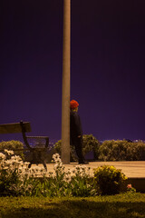 Mujer usando un beani rojo parada apoyada en un poste de eléctrico de perfil, día de invierno, está en un parque de noche en la cima de la ciudad