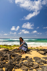 an African American woman wearing sunglasses with long sister locks relaxing at Sandy Beach with rocks covered in green algae, blue ocean water, crashing waves blue sky and clouds in Honolulu Hawaii