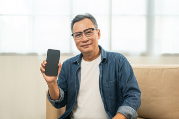Handsome asian mature old man using smartphone on sofa in living room at home. Happy Portrait of cheerful smiling senior asian man holding cell phone. Mature People and lifestyle