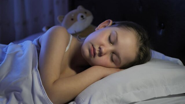 A cute 8-year-old Caucasian girl sleeps calmly and carefree in her bed on white pillows with her palms under her cheeks. Comfortable sleep for the child in good conditions. A healthy child sleeps well