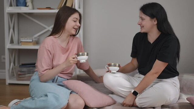 Tea drinking on the floor in the living room. Two girls talk, laugh and hold porcelain tea cups in their hands while sitting on the floor on a blanket in the room of a modern house