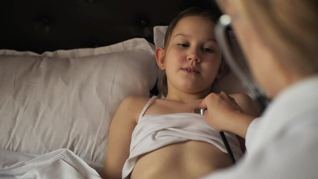 A female pediatrician uses a stethoscope to listen to the breathing of an 8 year old girl lying in bed and sick with a cold. A family doctor uses a stethoscope to listen to noises in a girls bronchi.