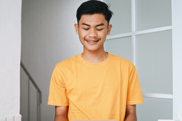 Young Asian man with braces looking down with happy expression against front door of the house