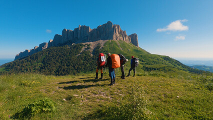 Four hikers with backpacks and trekking poles stand in front of a powerful detached rock. Sunny day...